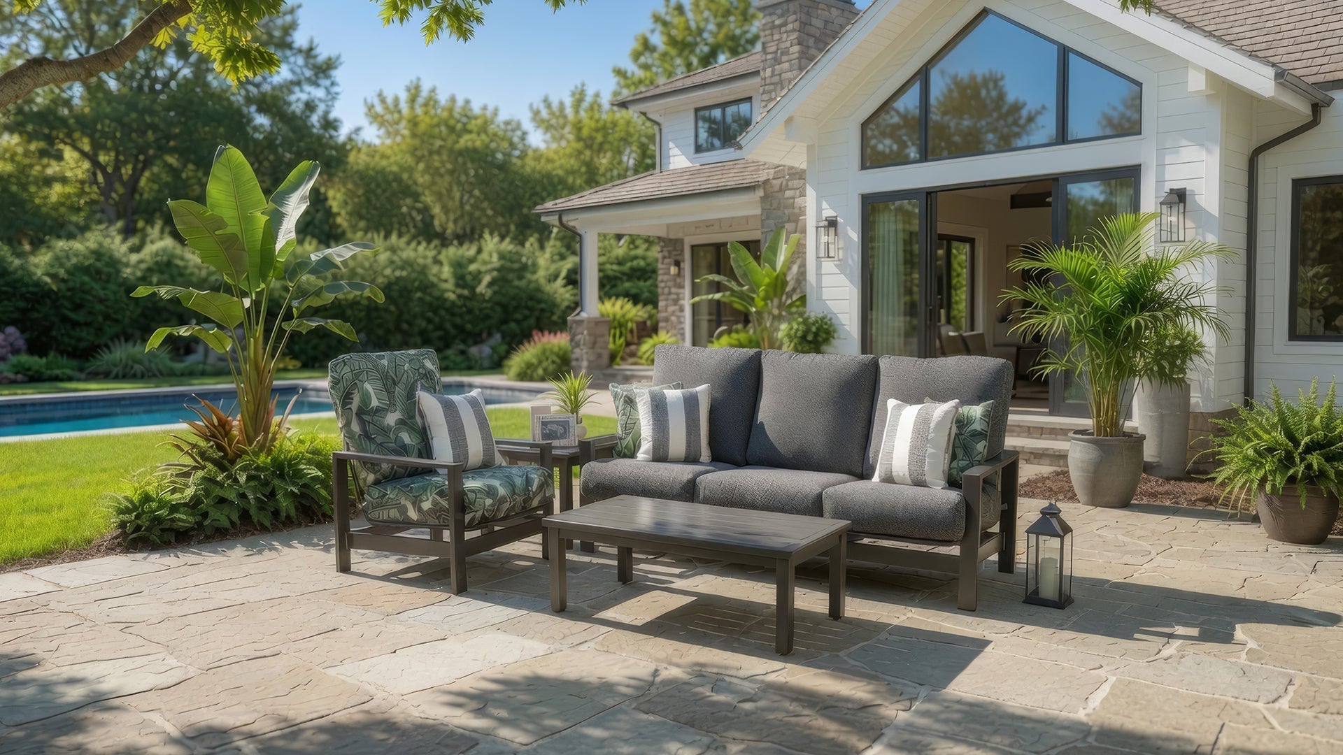 Outdoor seating set shown on residential rear patio with large square stone pavers. Outdoor seating set contains cushioned aluminum sofa with four toss pillows, cushioned club chair with toss pillow, square end table and rectangular cocktail table.